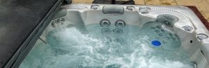 Close-up of a bubbling hot tub filled with water. The hot tub has multiple jets and built-in seats, with a partially open gray cover. A blue plastic ball floats on the water's surface. The surrounding area is a stone or concrete patio.