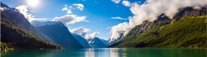 Panoramic view of a serene fjord with clear blue water surrounded by green, forested mountains. Snow-capped peaks are visible in the distance under a bright blue sky with scattered clouds. Sunlight gently illuminates the landscape.