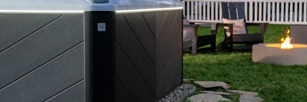 A Sundance Spas hot tub with a glowing edge sits in a backyard with green grass. Nearby, two dark chairs face a round fire pit with a small flame. In the background, a white picket fence is partially visible, offering an aura of excellence and relaxation.