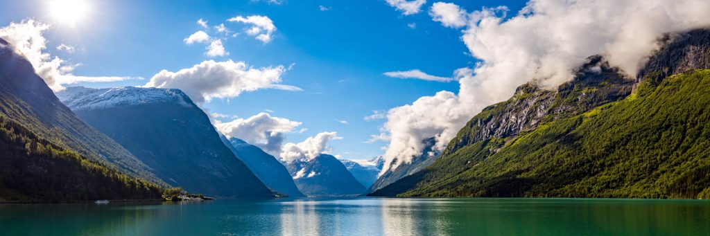A serene lake surrounded by lush green mountains under a bright blue sky with scattered clouds. Snow-capped peaks are visible in the distance, reflecting in the calm water, creating a picturesque and tranquil landscape, embodying Eco-Friendly Luxury akin to Earth Spas.