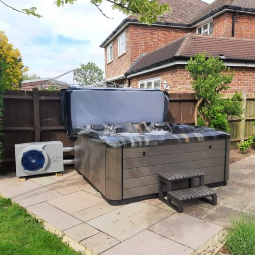 A gray hot tub with an open lid sits on a paved patio area beside a brick house. A small set of steps leads up to the tub. Behind it, there is a wooden fence and a white air conditioning unit by Mariana. The surroundings include a few plants and shrubs, creating a cozy retreat.