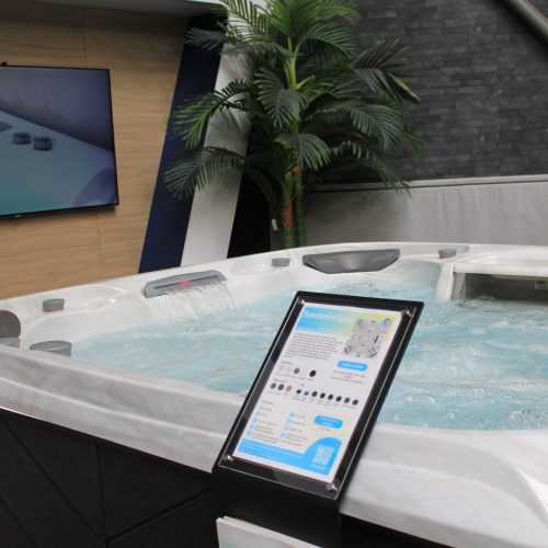A modern hot tub filled with water is displayed in an indoor showroom. There's a control panel stand in the foreground and a large TV screen on the wall showing a close-up of the hot tub's features, highlighting its award-winning design from the UK Pool & Spa Awards. A potted palm tree is nearby.