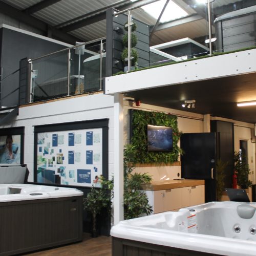 Showroom featuring multiple outdoor hot tubs. There are plants and informational displays visible, along with modern glass railings leading to an upper mezzanine level. Shelving with various products is seen in the background, illuminated by overhead lights.